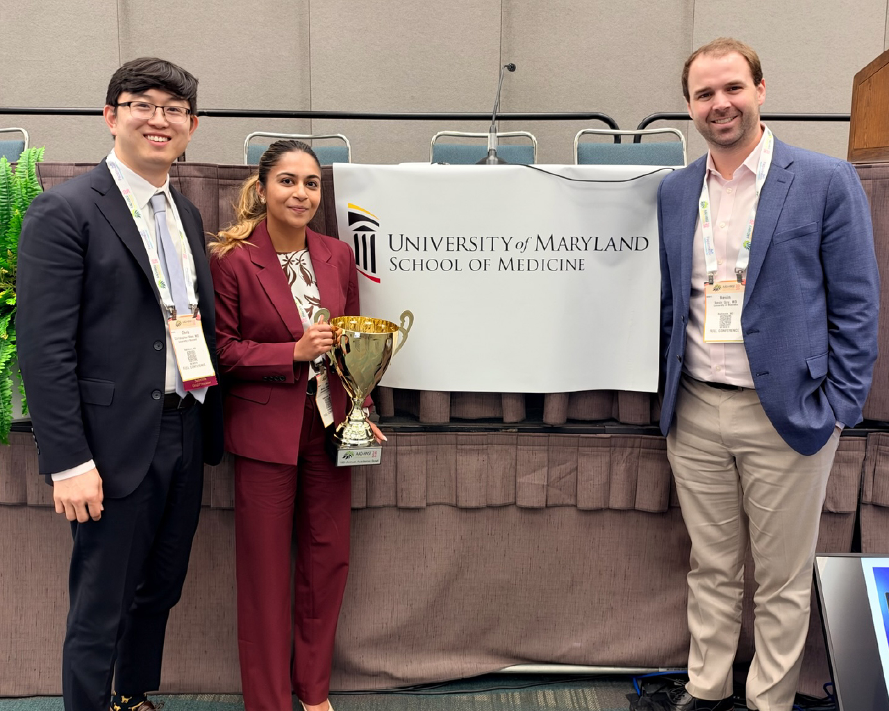 The winning team of the 2026 Academic Bowl from the University of Maryland School of Medicine. Two men and one woman stand indoors on a stage, proudly holding an Academic Bowl trophy.
