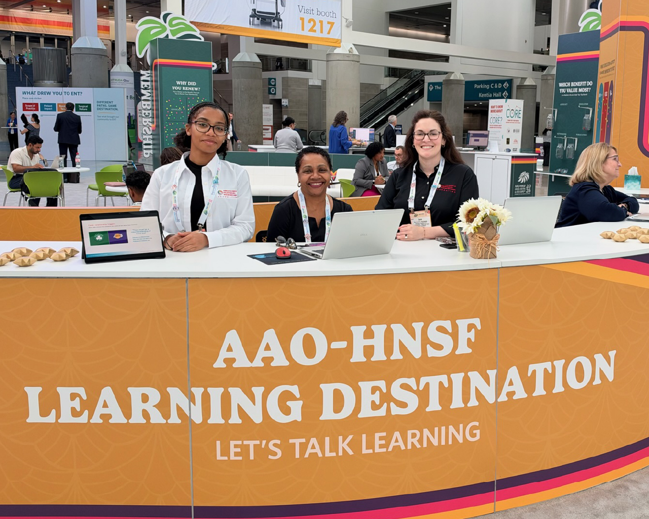 Staff members seated at the vibrant orange and yellow AAO-HNSF Learning Destination booth in a busy convention center lobby.