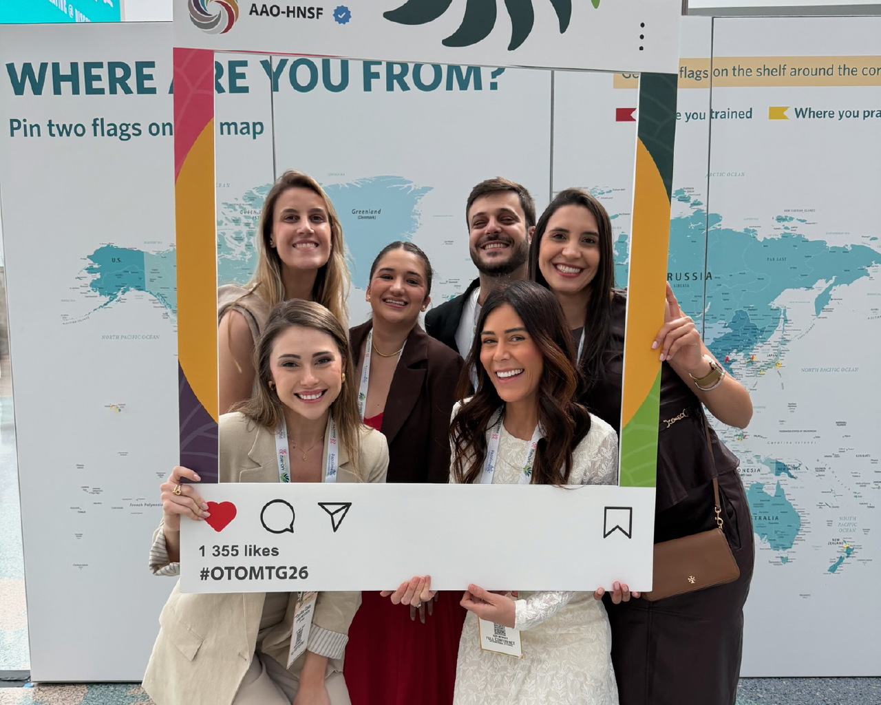 A group of six smiling attendees posing together behind a large, social-media-style photo frame cutout.