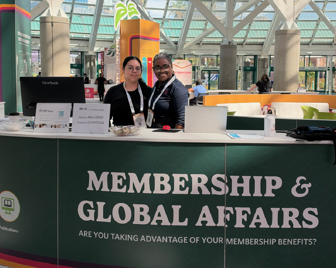 Two staff members sitting behind a wide dark green counter labeled Membership & Global Affairs in a spacious lobby area.